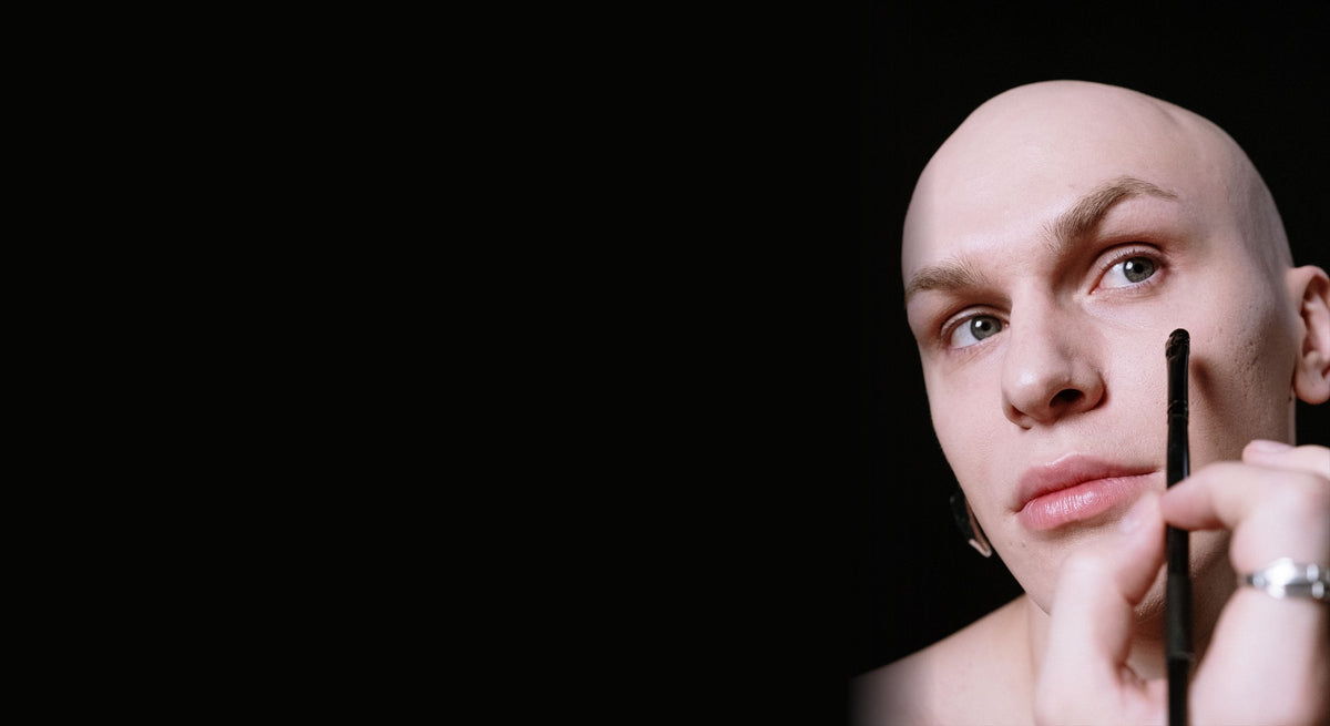 Man with a clean, even complexion holding a makeup brush to his cheek against a black background